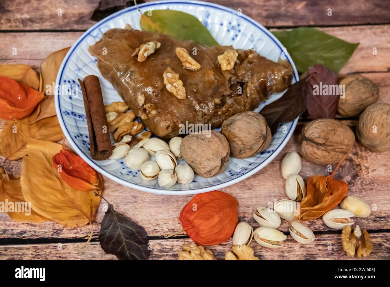 Turkish Baklava sweet cake on the plate, decorated with fresh pistachios, nuts, lemon, apple, vanilla and dry autumn leaves, on wooden massive table Stock Photo