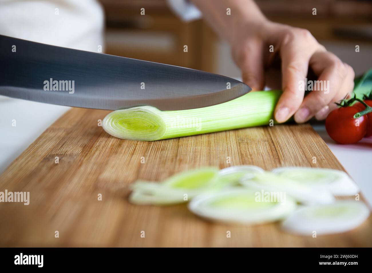 Close-up of hand slicing leek with chef's knife on a bamboo cutting board, tomatoes in background Stock Photo