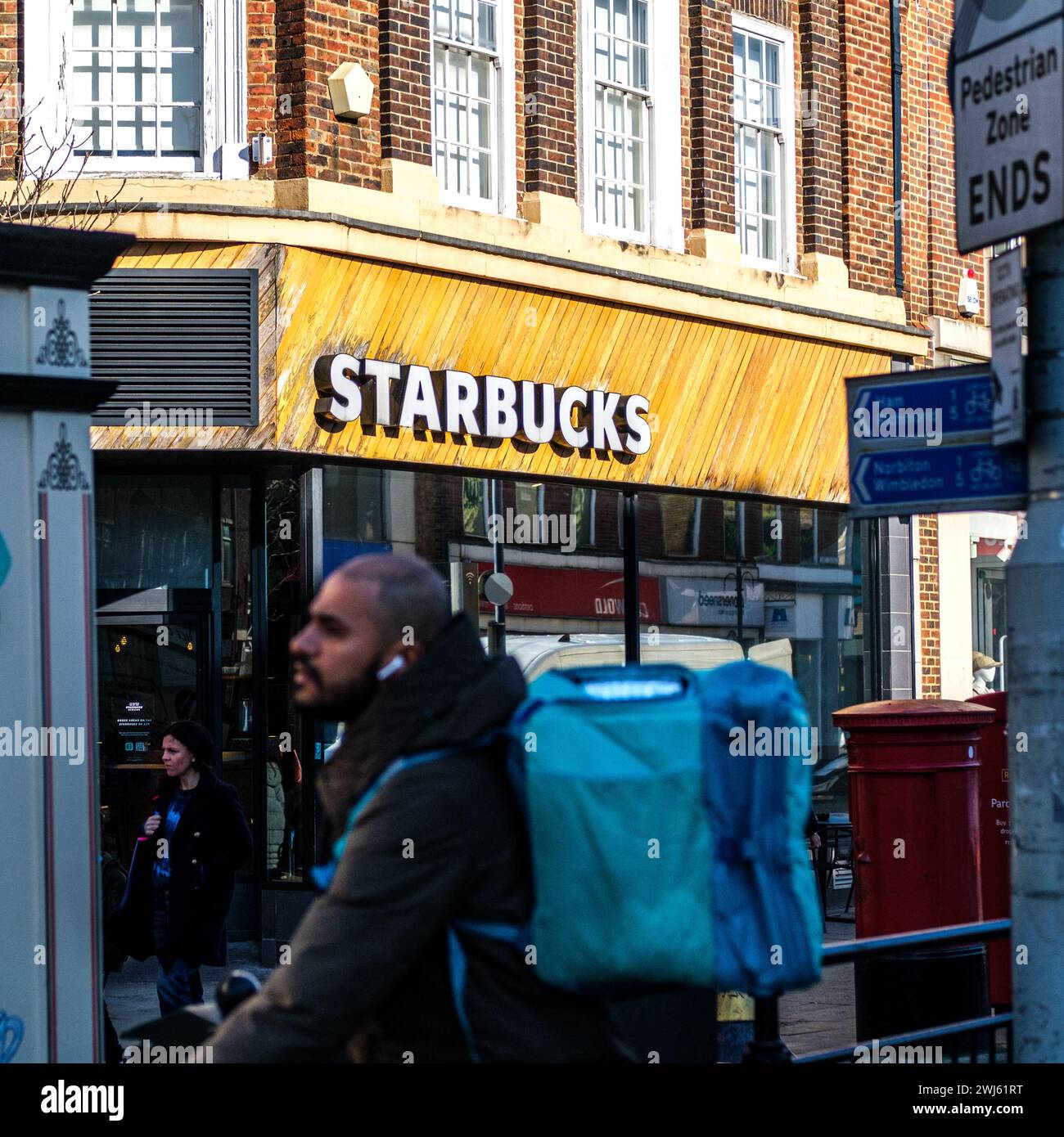 Kingston-Upon-Thames, London UK, February 12 2024, Starbucks Coffee Shop Lit By Morning Sunlight With Deliveroo Rider Passing In Foreground Stock Photo