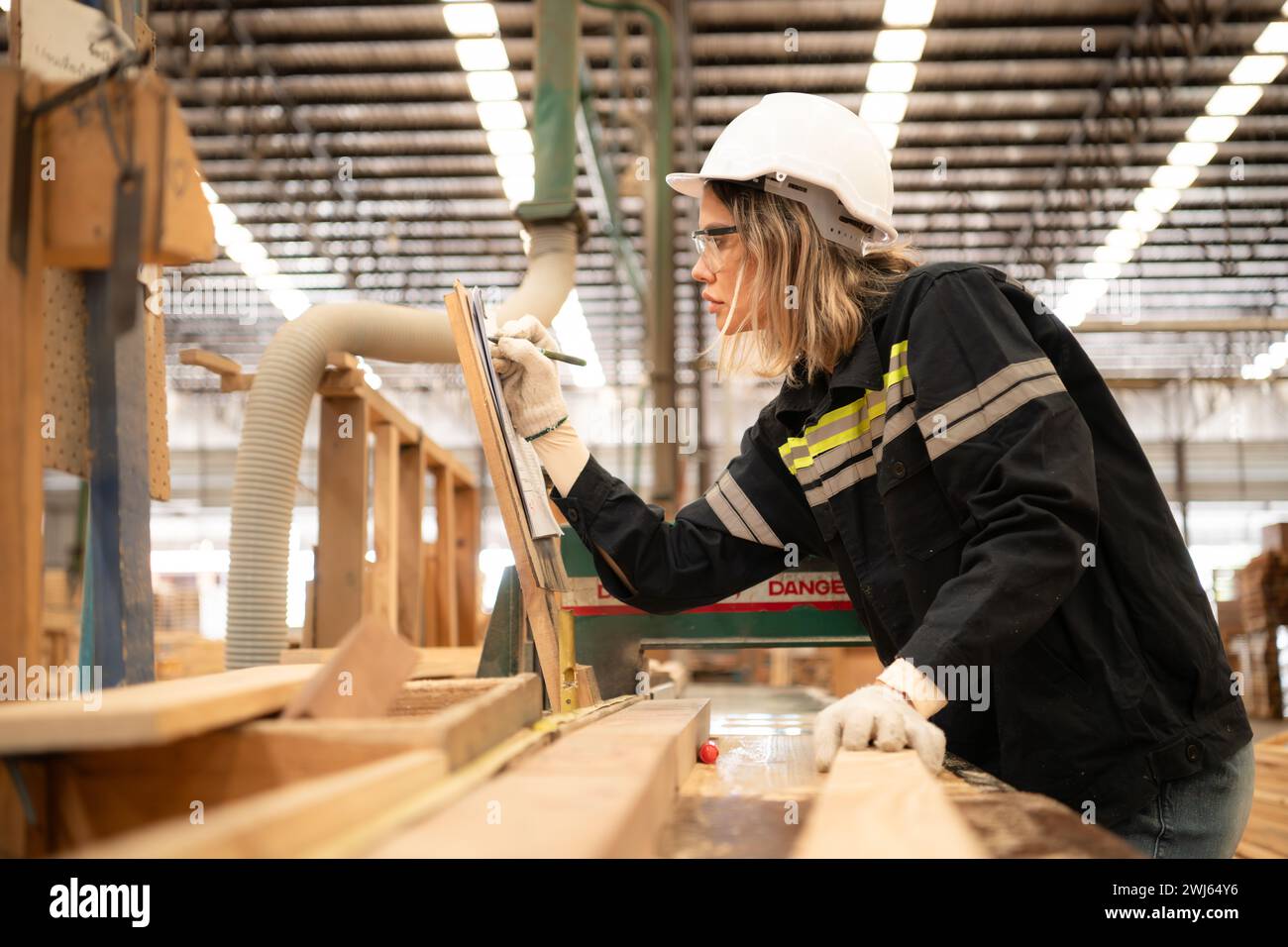 Female carpenter working in a woodworking factory, She is wearing a safety helmet and glasses. Checking the neatness of the plan Stock Photo