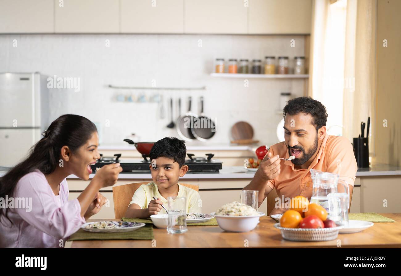 Happy parents with child eating lunch together on dining table at home - concept of weekend relaxation, family bonding and parenthood Stock Photo