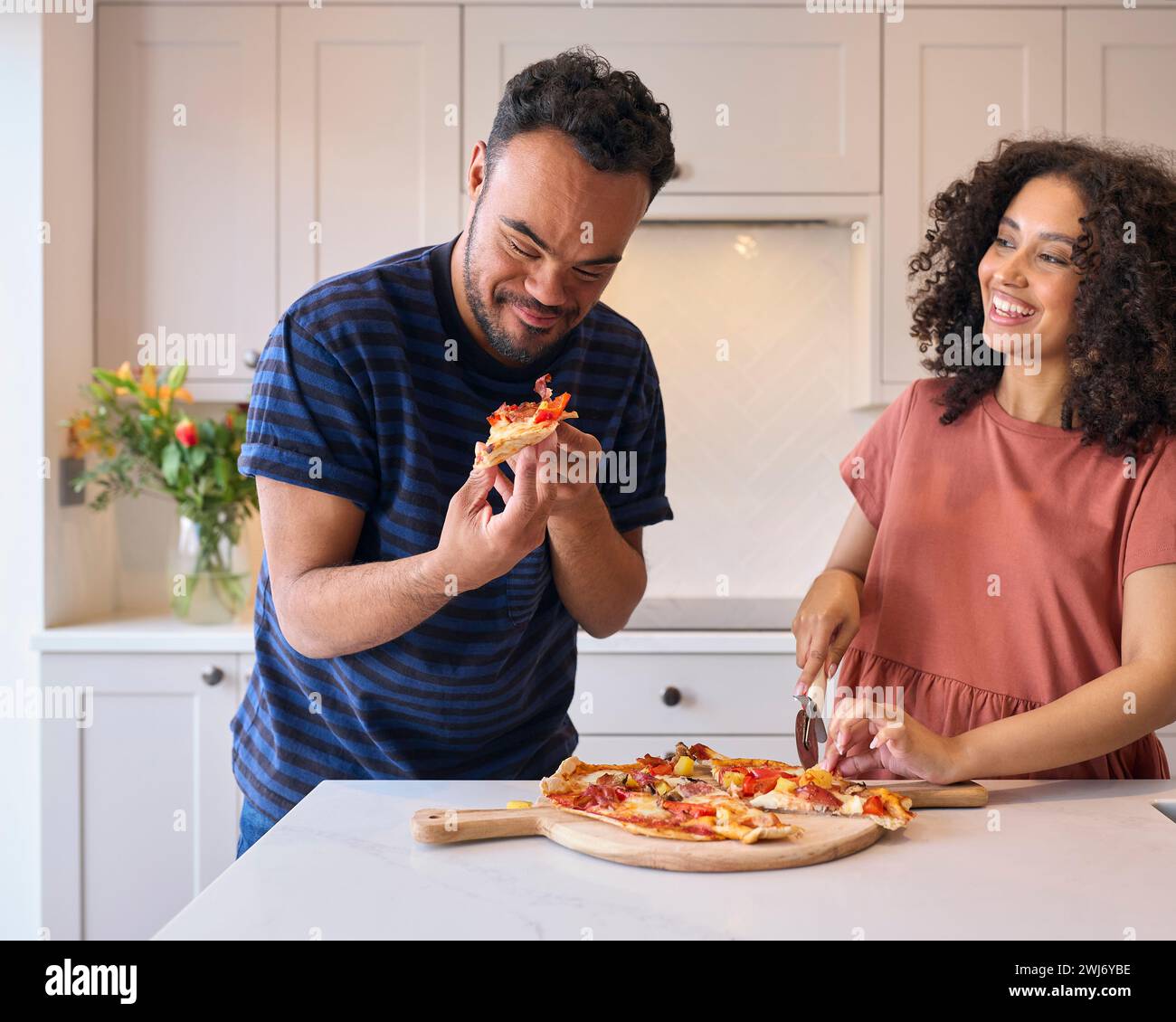 Couple At Home With Man With Down Syndrome And Woman Eating Homemade Pizza In Kitchen Together Stock Photo