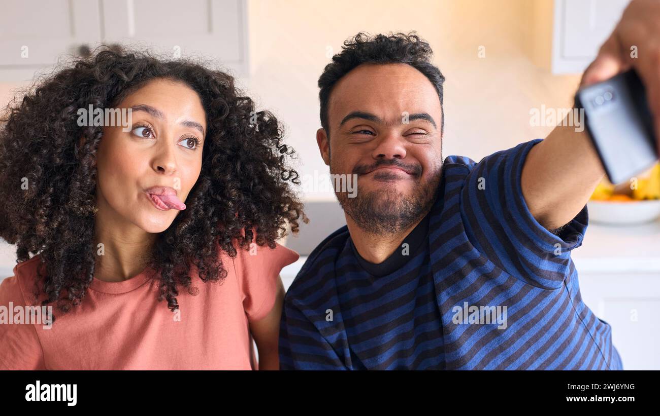 Couple At Home With Man With Down Syndrome And Woman Posing For Selfie On Mobile Phone Stock Photo