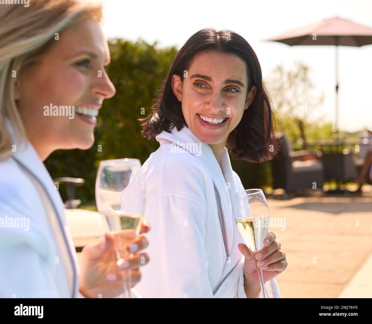 Two Mature Female Friends Wearing Robes Outdoors By Pool Drinking
