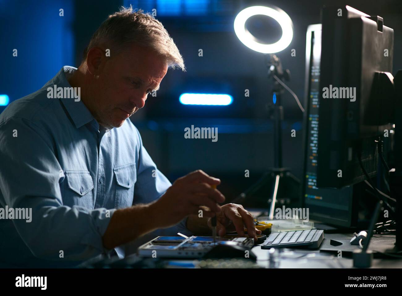 Mature Male Electronics Expert Repairing Laptop In Workshop With Low Key Lighting Stock Photo