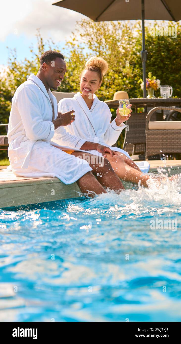 Loving Couple Wearing Robes Outdoors Sitting With Drinks Splashing Feet In Swimming Pool On Spa Day Stock Photo