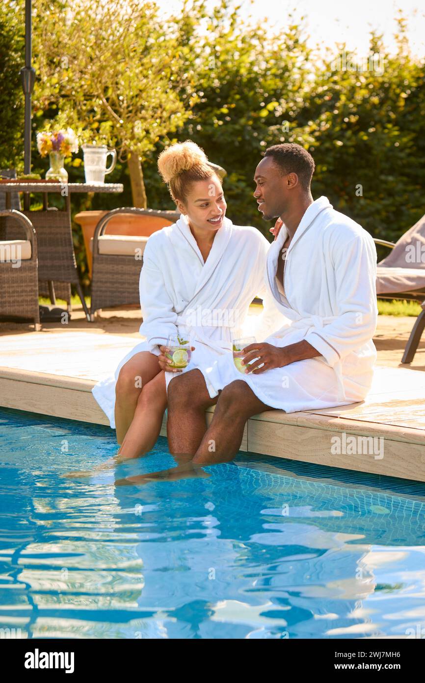 Couple Wearing Robes Outdoors Sitting With Drinks With Feet In Swimming Pool On Spa Day Stock Photo