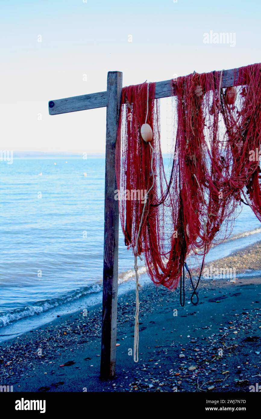 Red fishing net with buoys on wooden post at beach, calm sea, clear sky backdrop. Stock Photo