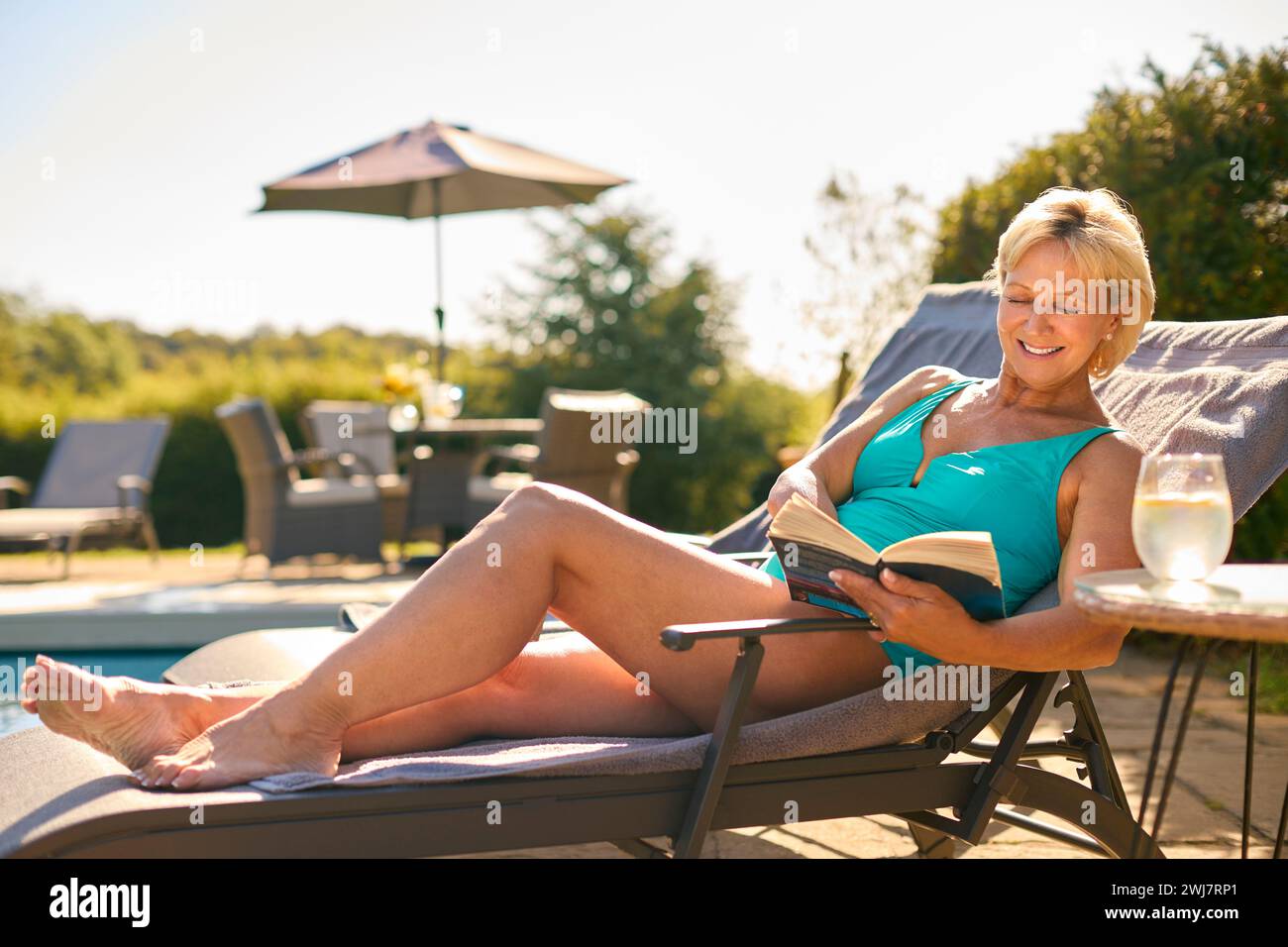 Senior Woman On Holiday Wearing Swimming Costume Reading Book Relaxing By Hotel Swimming Pool Stock Photo