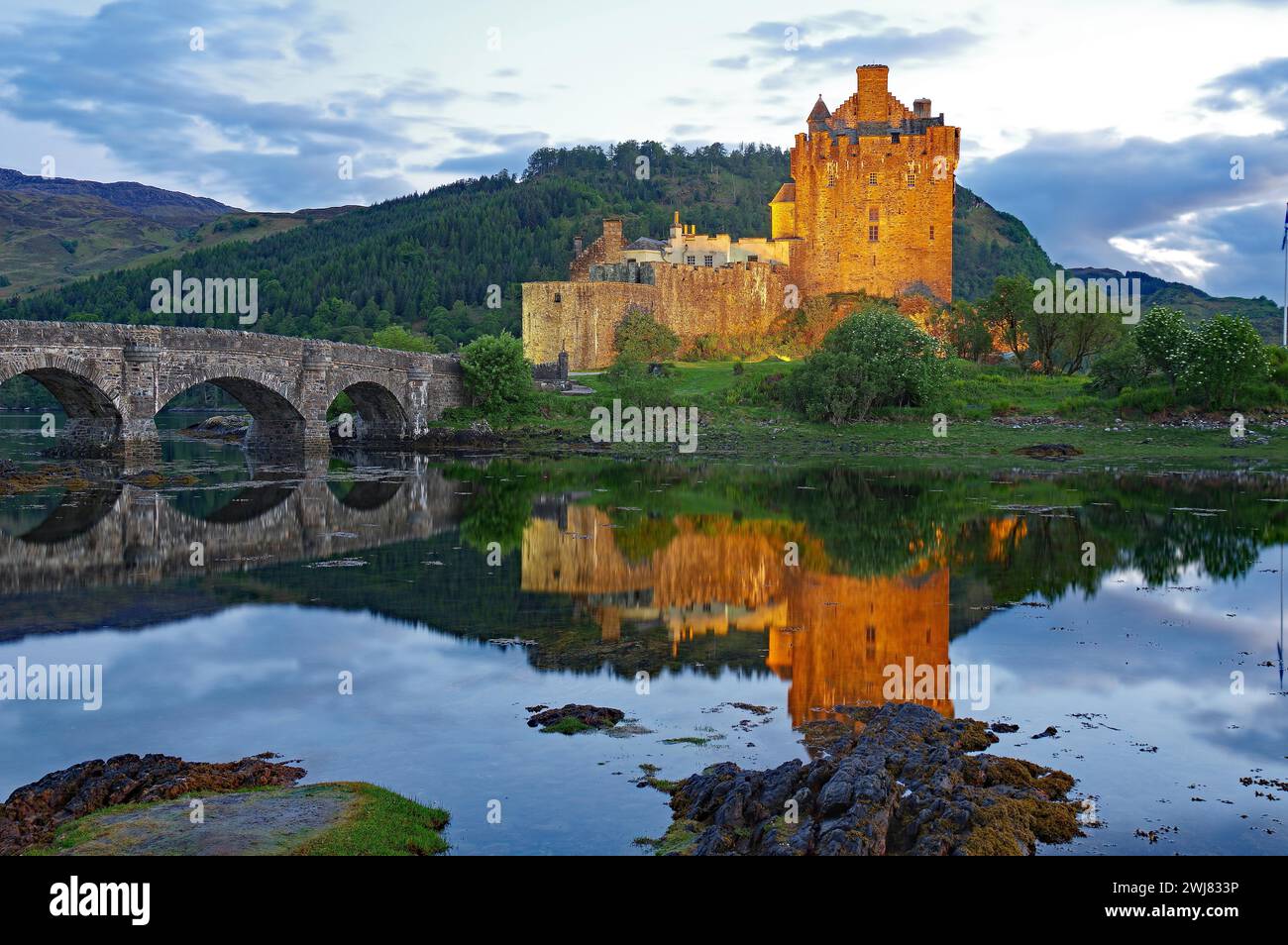 The illuminated Eilean Donan Castle is reflected in the water on a calm evening, old stone bridge, film location for James Bond, Highlander, Rob Roy Stock Photo