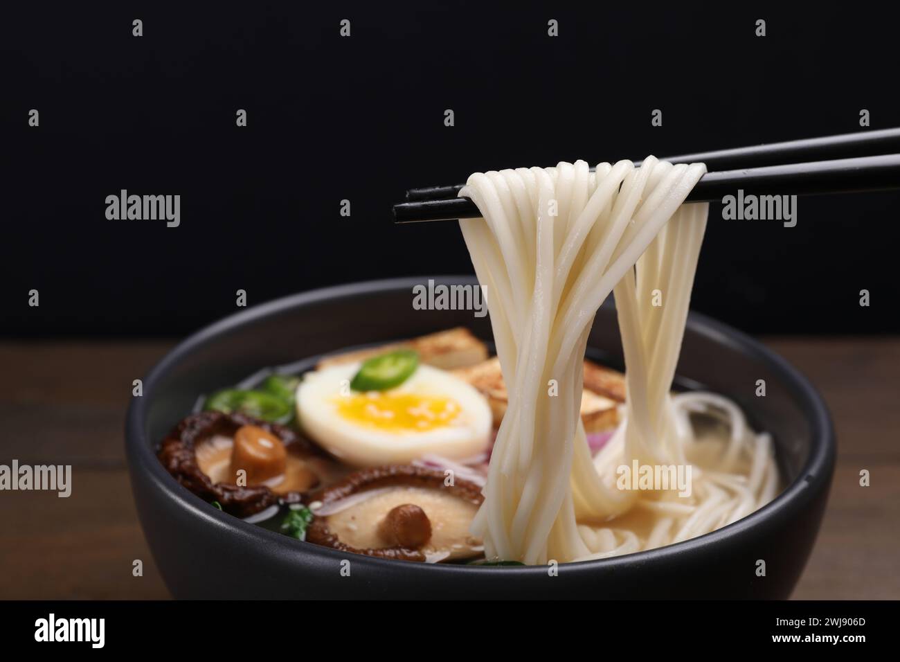 Eating delicious vegetarian ramen with chopsticks at table against black background, closeup Stock Photo