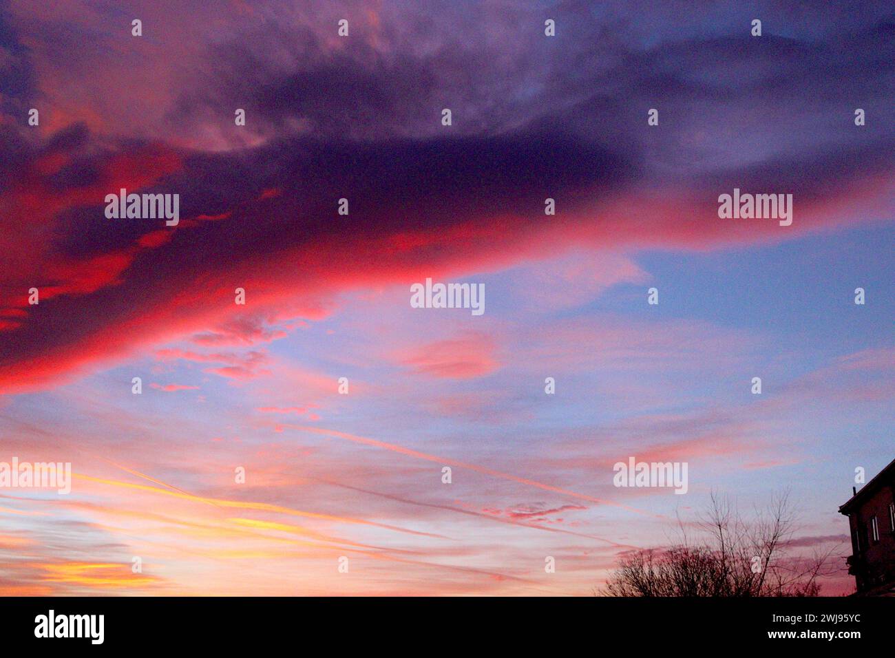 Spectacular surreal sunset with aircraft vapour trails across the sky catching rays of evening light, contrast against multicoloured clouds. Stock Photo