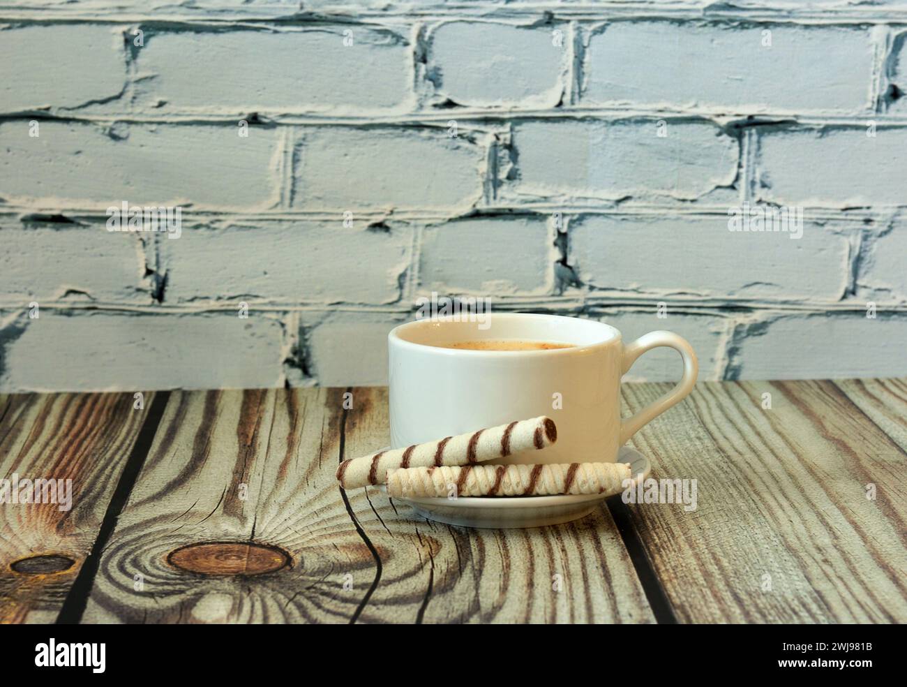 A cup of hot fresh cappuccino on a saucer with two wafer rolls on a light wooden table. Close-up. Stock Photo