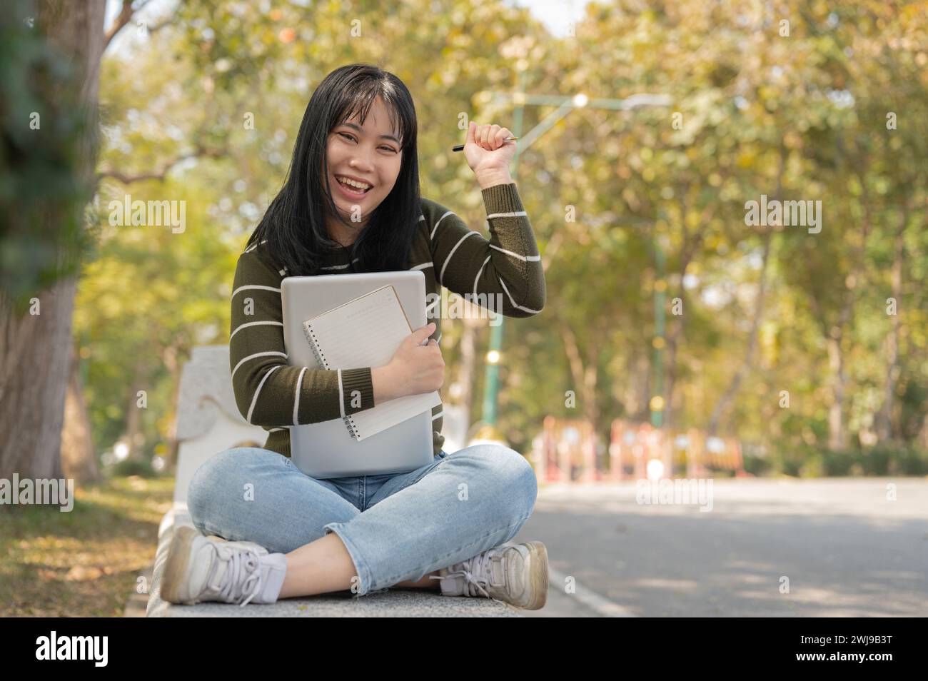 A cheerful young Asian female college student is sitting on bench in a ...