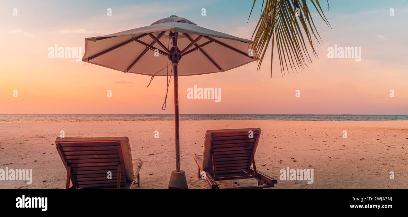 Beautiful tropical sunset scenery, two sun beds, loungers, umbrella under palm tree. White sand, sea view with horizon, colorful twilight sky, calm Stock Photo