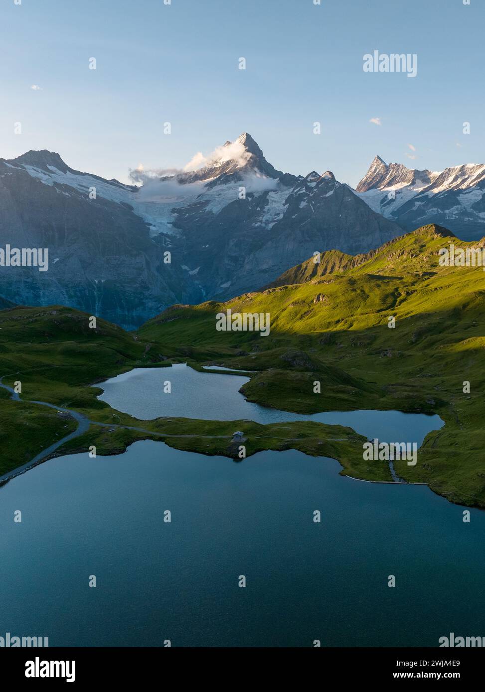 Tranquil alpine lake nestled among rolling green hills with rugged mountains in the backdrop at sunset. Stock Photo