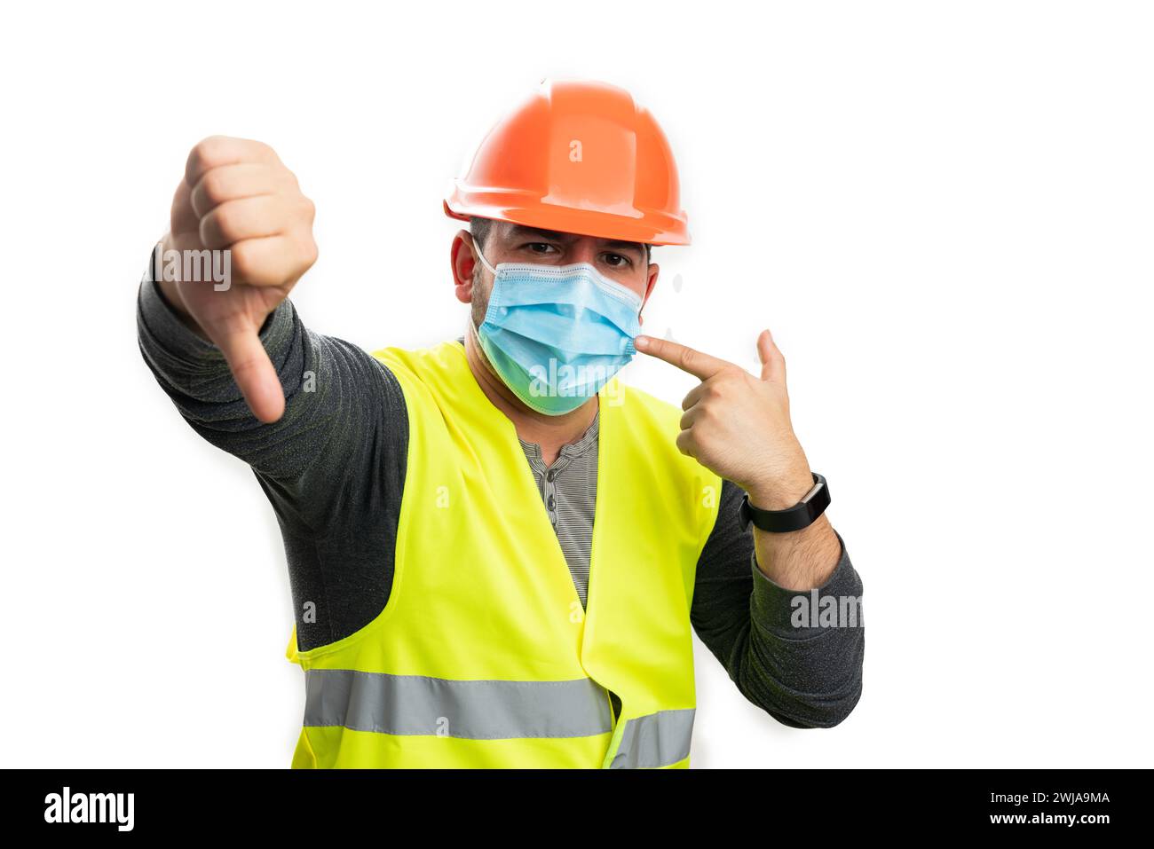 Constructor man wearing fluorescent vest and hardhat pointing index finger at medical or surgical mask making thumb-down dislike gesture as pandemic c Stock Photo