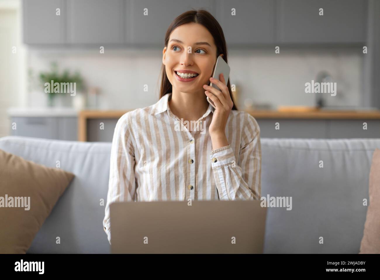 Happy young woman talking on phone and using laptop at home Stock Photo