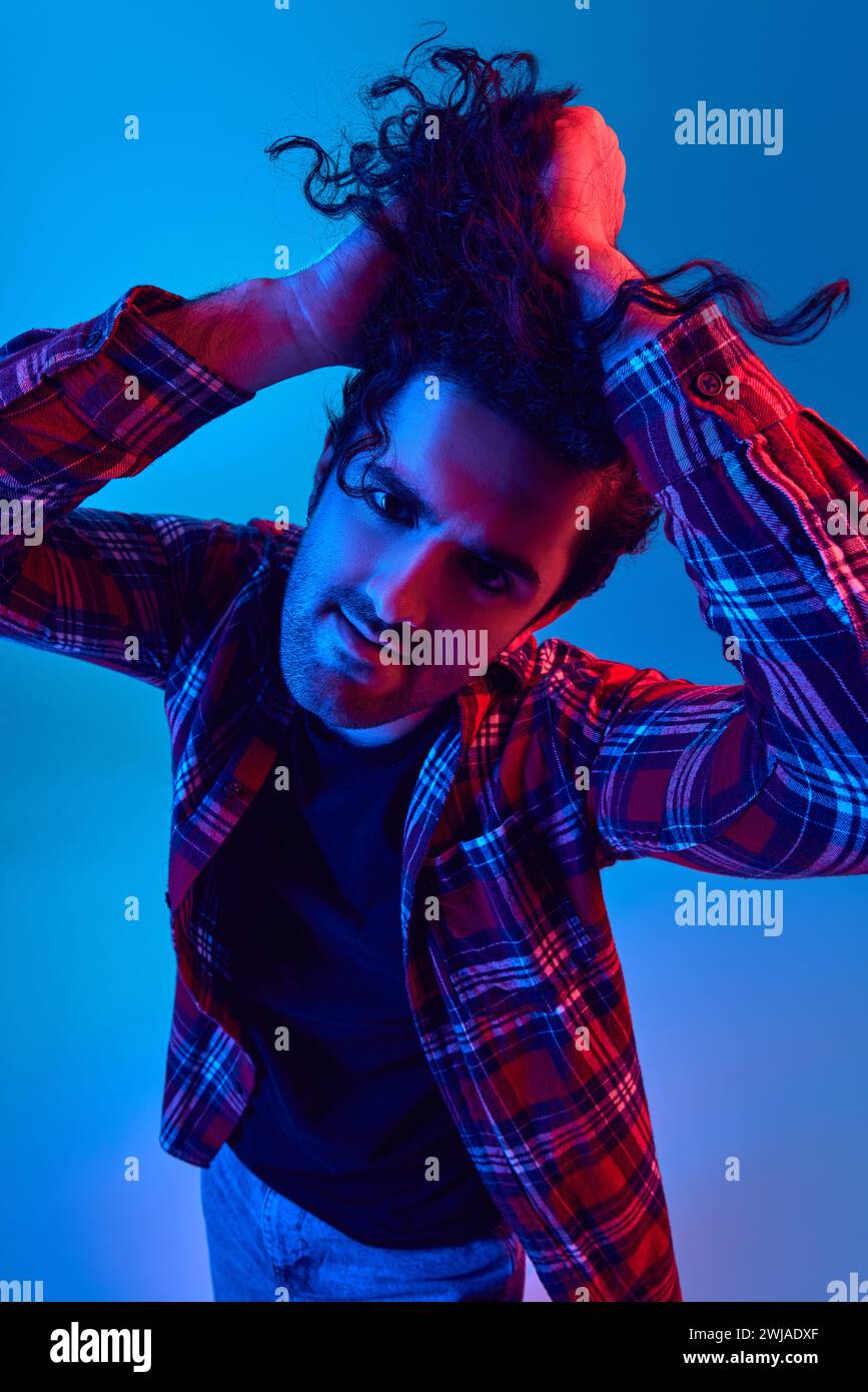 Crazy, queer. Young guy with curly hair, unshaved face, wearing checkered shirt and looking at camera against blue background in neon light Stock Photo