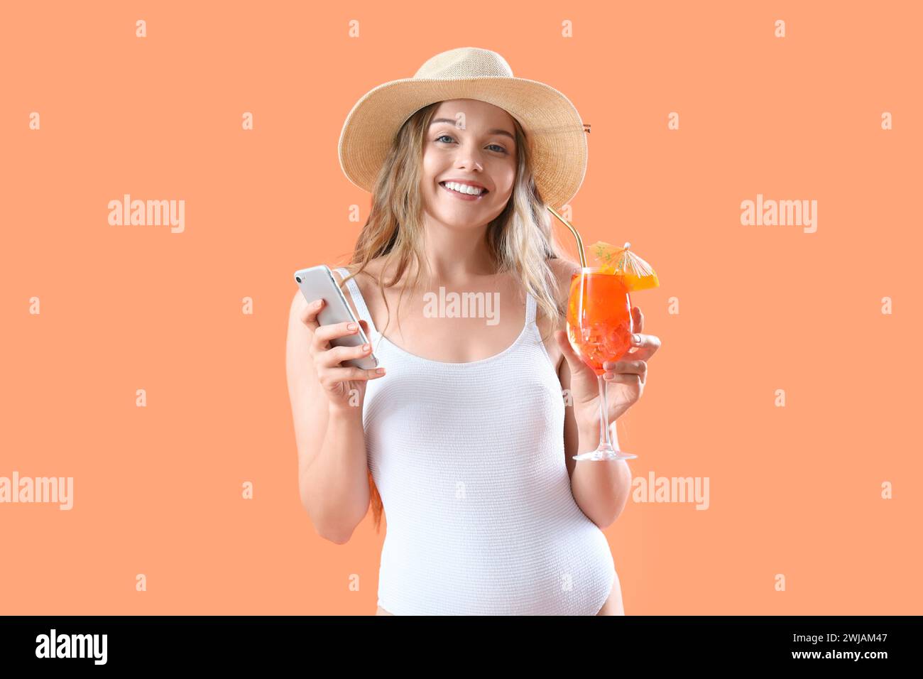 Beautiful young woman in swimsuit with glass of tasty aperol spritz and mobile phone on orange background Stock Photo