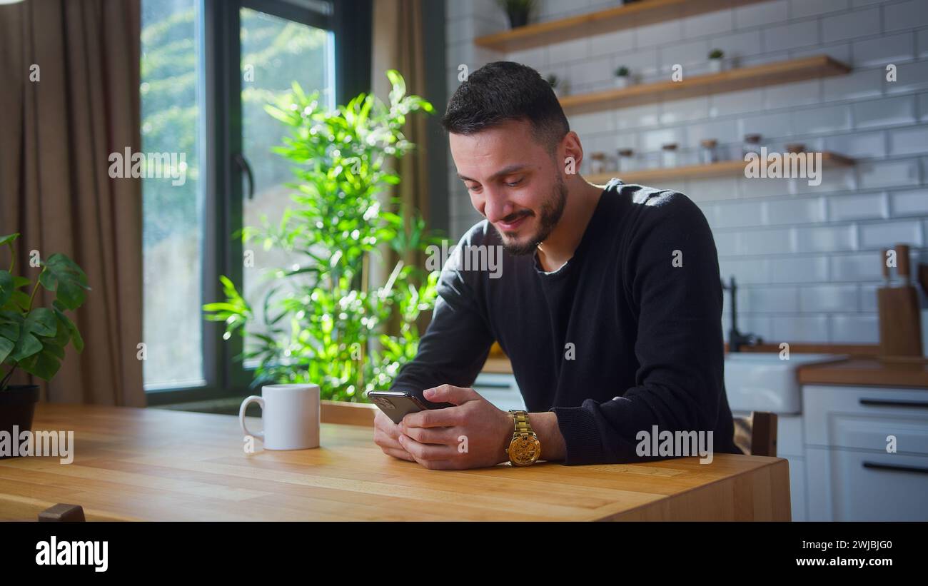 Smiling man sit in kitchen surfing internet on mobile phone, reading media news, scrolling social networks, using mobile applications on smartphone at Stock Photo