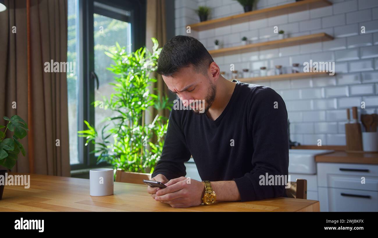 man sit in kitchen surfing internet on mobile phone, reading media news, scrolling social networks, using mobile applications on smartphone at home Stock Photo