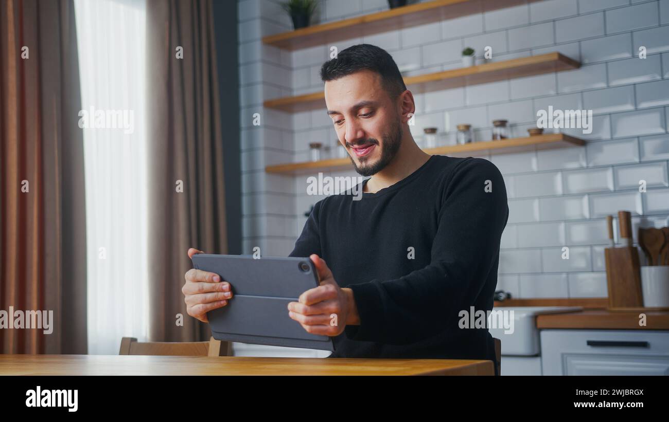 Adult man playing a video game on a tablet computer while sitting in kitchen at home. Excited concentrated man engrossed in interesting racing game, d Stock Photo