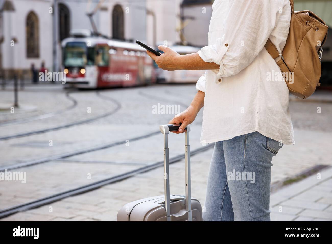 Woman tourist with suitcase and smart phone waiting for tram on street. Travel by public transportation in city Stock Photo