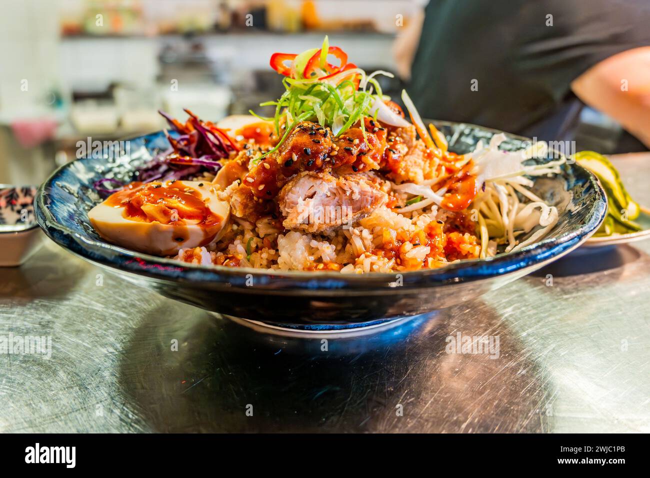 Sweet and sour asian bowl with chicken, shrimp, seed, egg and vegetable in blue bowl on table, part of man. Stock Photo