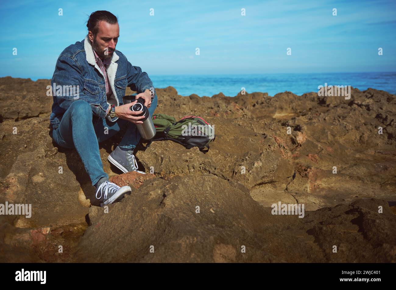 Caucasian bearded serious man, hiker, traveler, adventurer sitting on the rocky cliff by seam holding a stainless steel thermos with hot drink. People Stock Photo