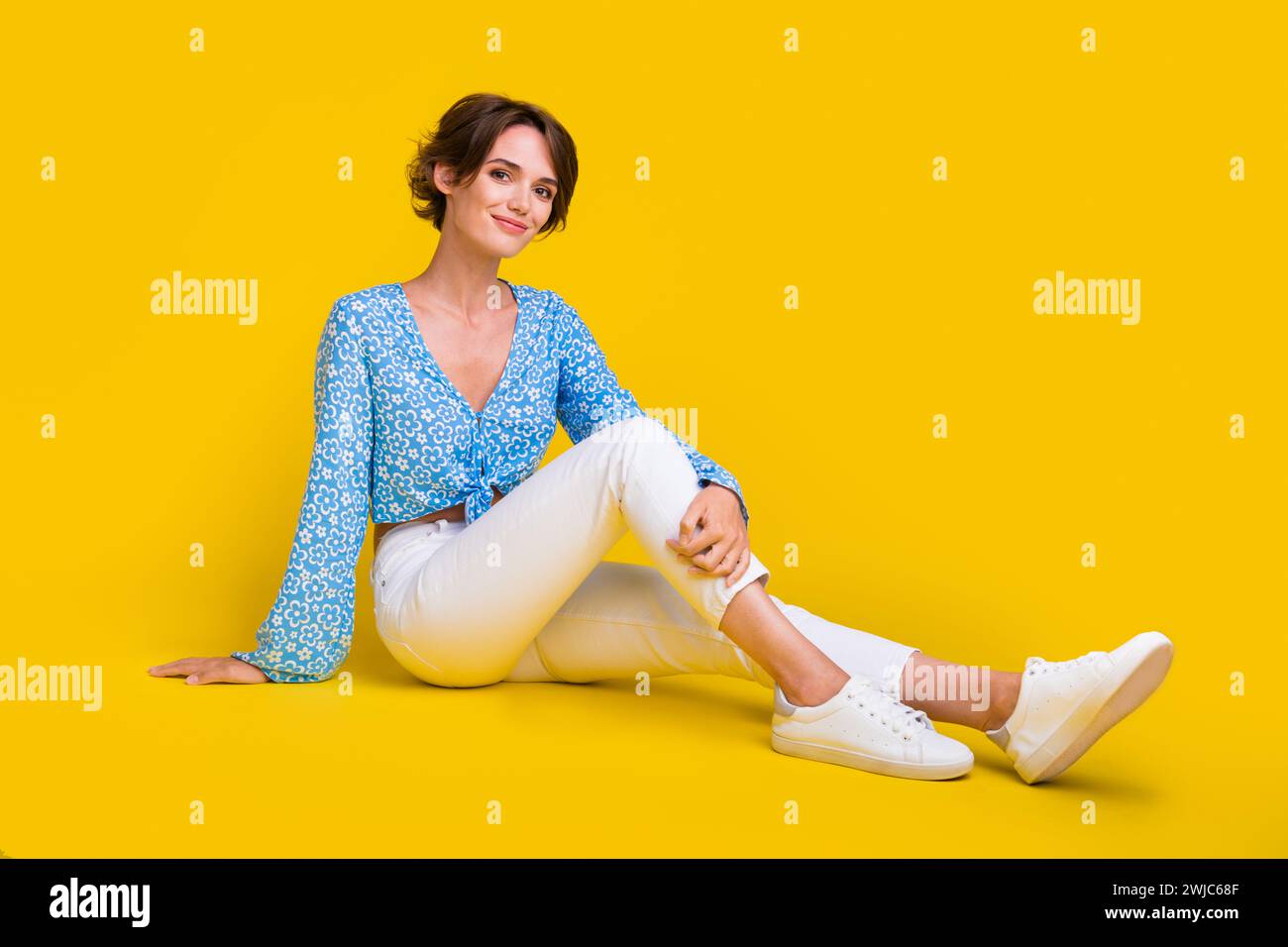 Full size photo of adorable gorgeous optimistic woman wear blue top white trousers sit on floor isolated on yellow color background Stock Photo