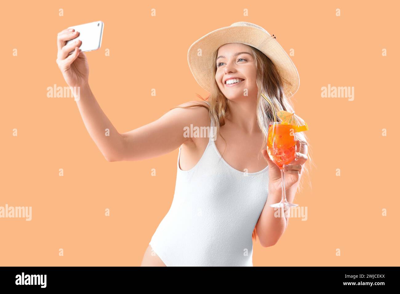 Beautiful young woman in swimsuit with glass of tasty aperol spritz taking selfie on orange background Stock Photo