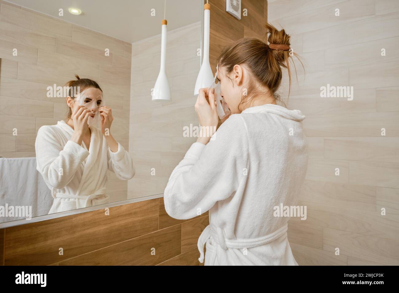 A woman in a white robe is applying skincare mask to her face, standing before a large mirror in a well-lit, contemporary bathroom Stock Photo