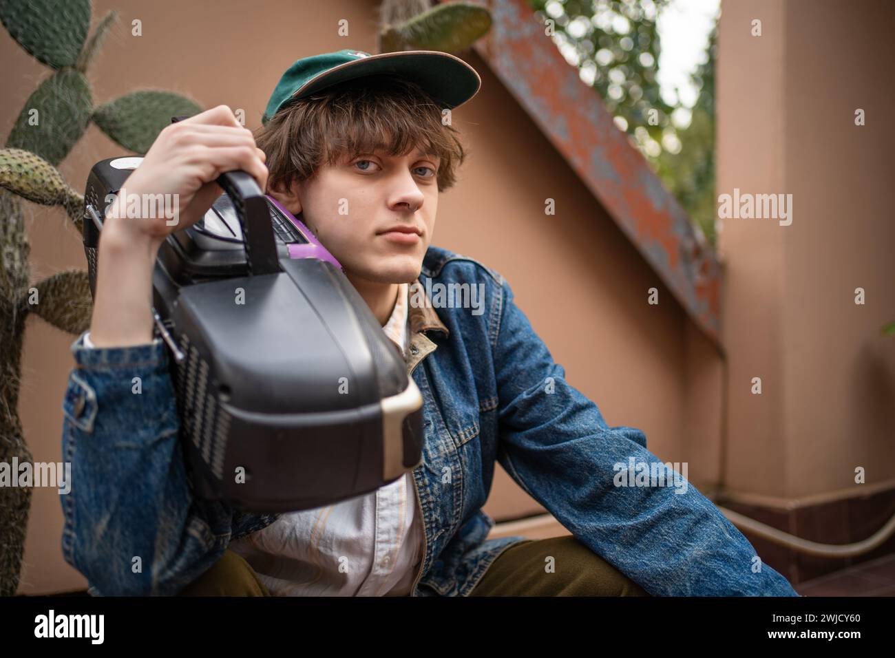 Portrait of young relaxed man looking at camera. His clothes and accessories reflect style of the 90s. Cassette player is nostalgic element that adds retro charm to photo. Stock Photo