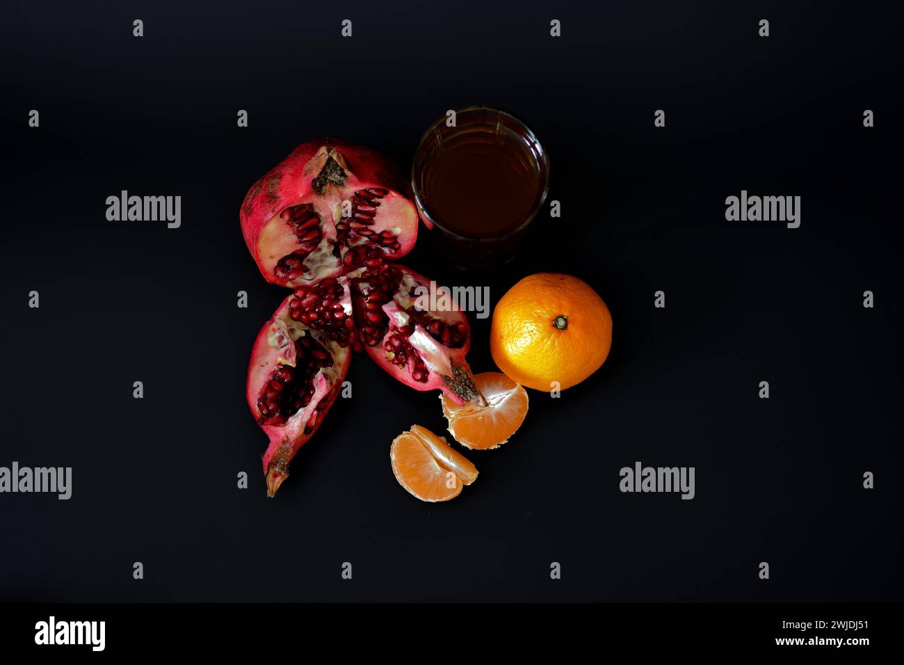 A tall glass of a mixture of fruit juices on a black background, a ripe tangerine and a broken pomegranate fruit with seeds. Close-up. Stock Photo