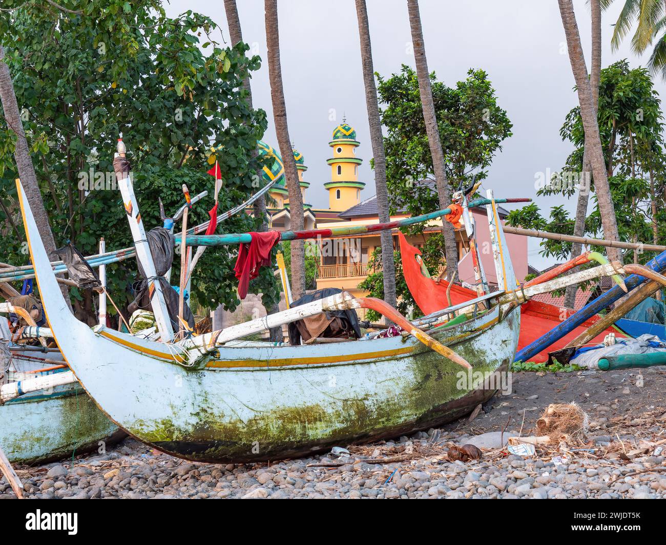 Traditional Indonesian fishing boats with outriggers at a stony beach in Medewi on the island of Bali in Indonesia. The Jami Masjid Al-Akmal mosque in Stock Photo