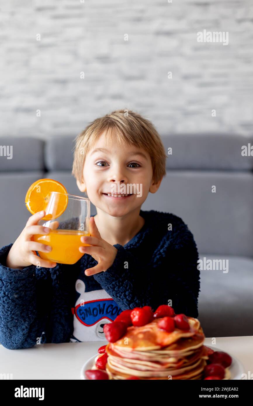 Sweet toddler child, boy, eating american pancakes with strawberries and bananas, topped with syrup and drinking fresh orange juice Stock Photo