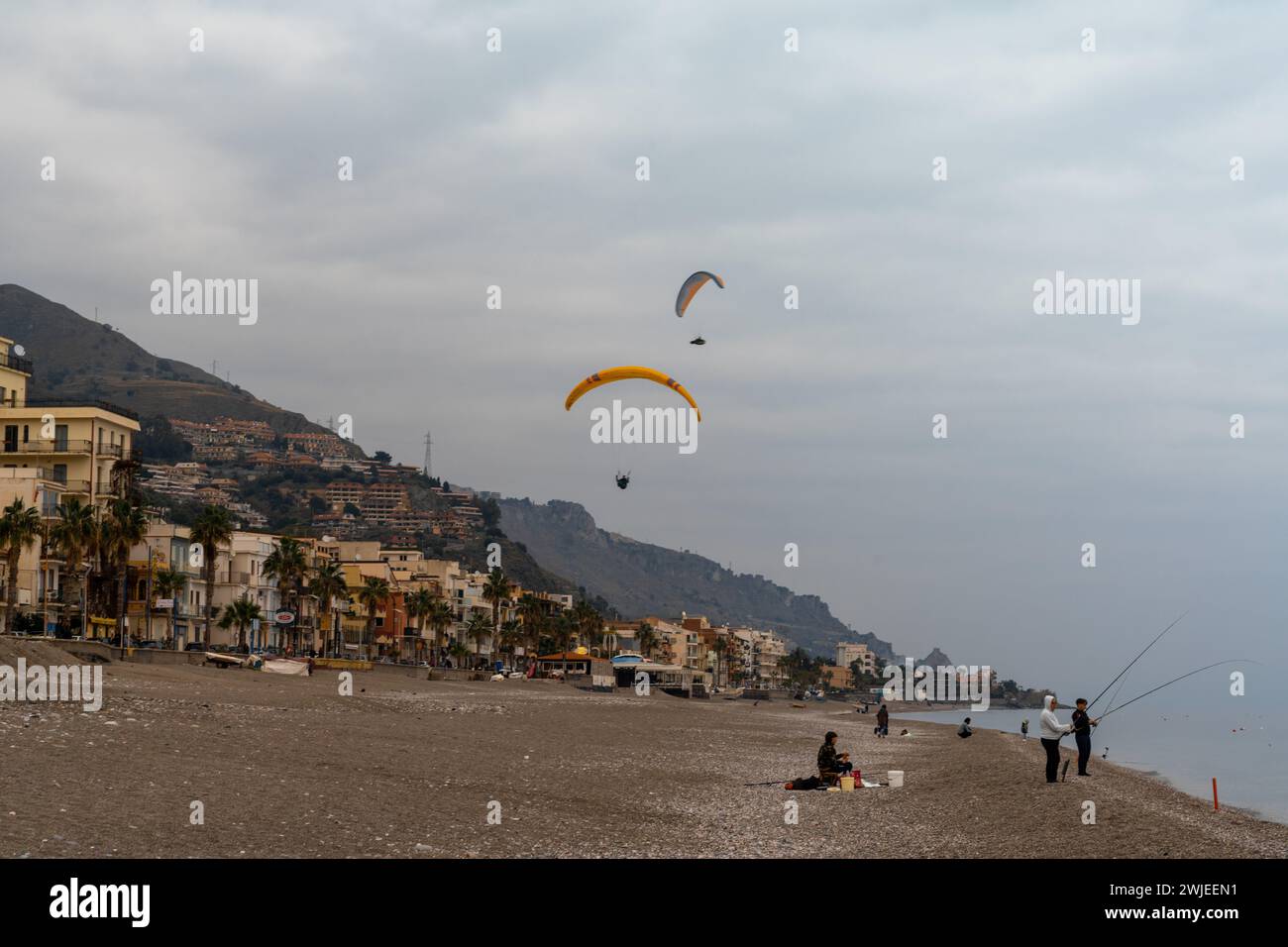 Letojanni, Italy - 29 January, 2023: fishermen on the beach at Letojanni with paragliders landing behind them after a flight from the Sicilian mountai Stock Photo