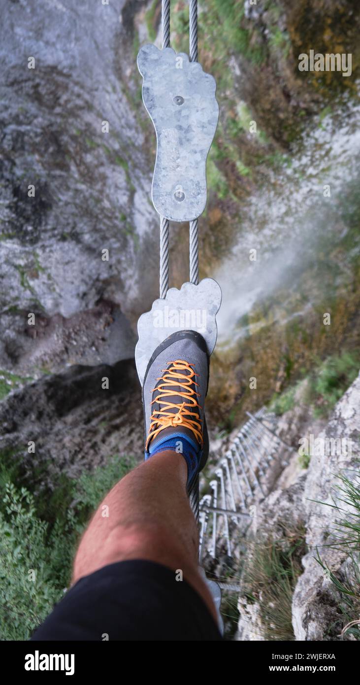 Tourist on foot shaped metal steps, on suspended bridge,over a waterfall, on via ferrata route in Italy, Europe. Summer adventure activities in the Do Stock Photo