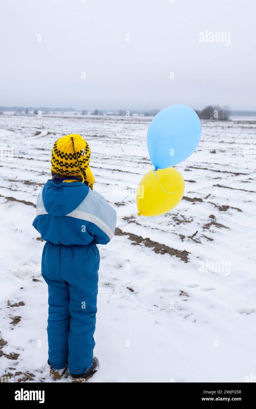 boy standing alone with his back on a snowy field with yellow and blue balloons. drawing attention to the war in Ukraine. stolen childhood, refugee pr Stock Photo