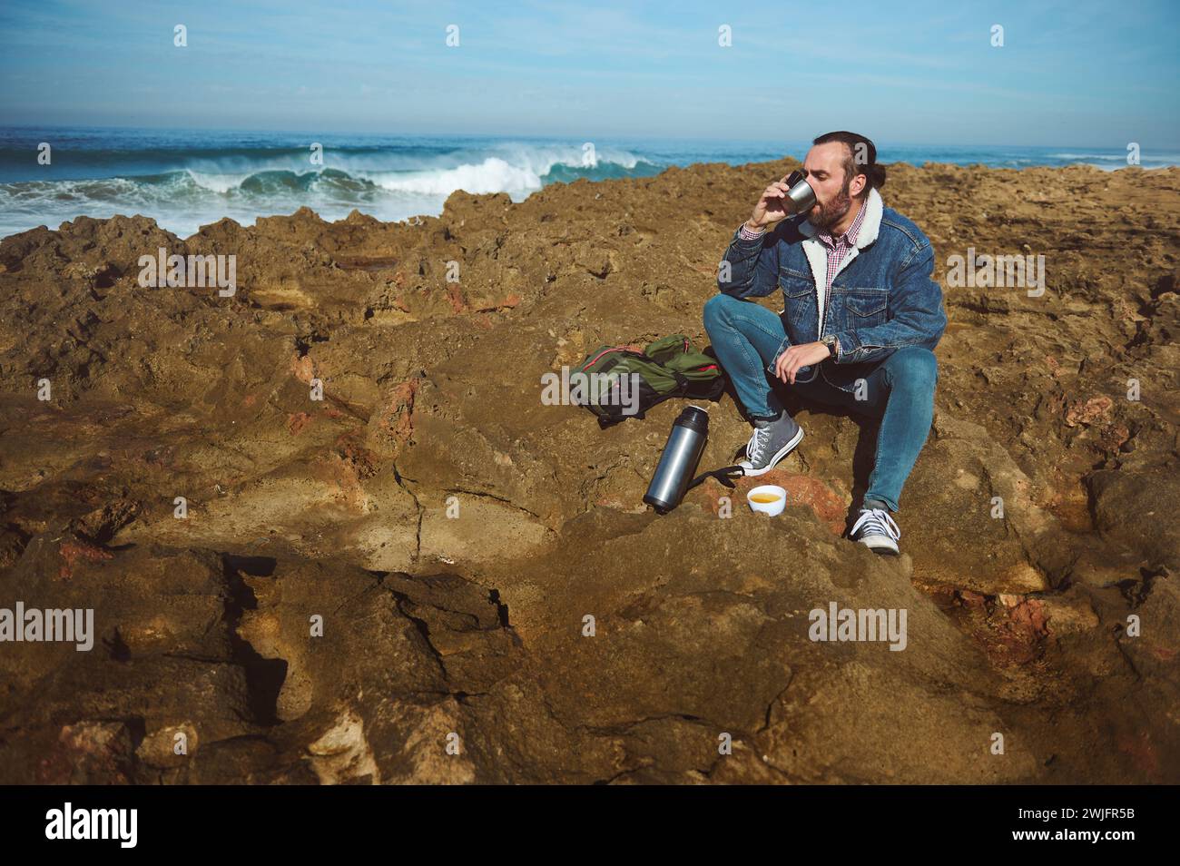 Portrait of young adult bearded man in casual denim clothes, in the beach, drinking hot tea outdoors from thermos. Hiking. Stock Photo