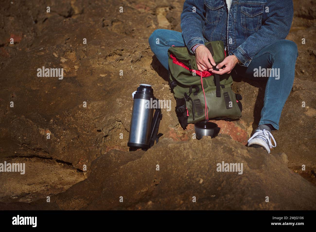 Close-up Caucasian bearded brutal male tourist traveler adventurer with backpack and thermos flask, sitting on the cliff by sea, enjoying his coffee b Stock Photo