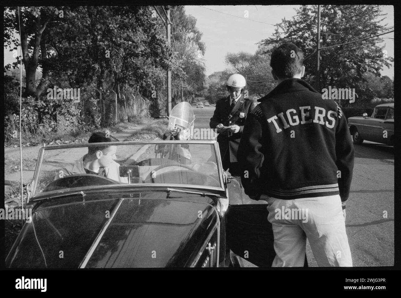 Teen driver Bill Kolb (in Tigers jacket with back to the camera) receives a traffic ticket from a police officer, White Plains, New York, circa 1958. (Photo by Charlotte Brooks/LOOK Magazine Collection/LOC) Stock Photo