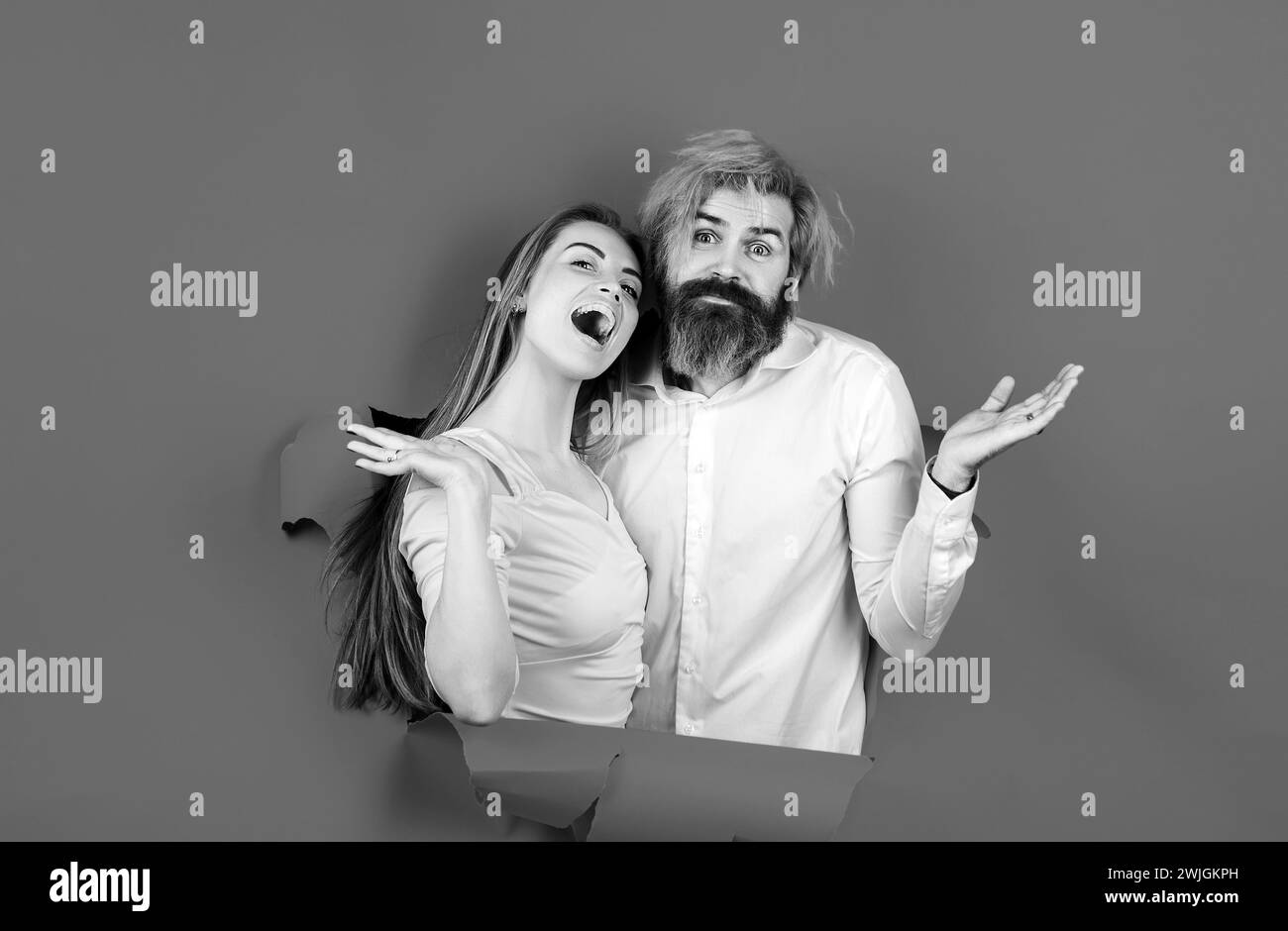 Surprised couple of bearded man and happy woman making hole in paper looking through hole. Advertising board, blue background. Couple through hole in Stock Photo