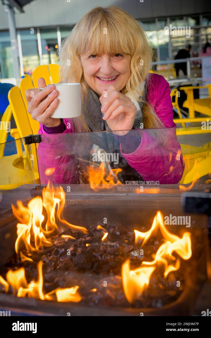 Woman enjoying mug of coffee outdoors by warm fire in winter Stock Photo