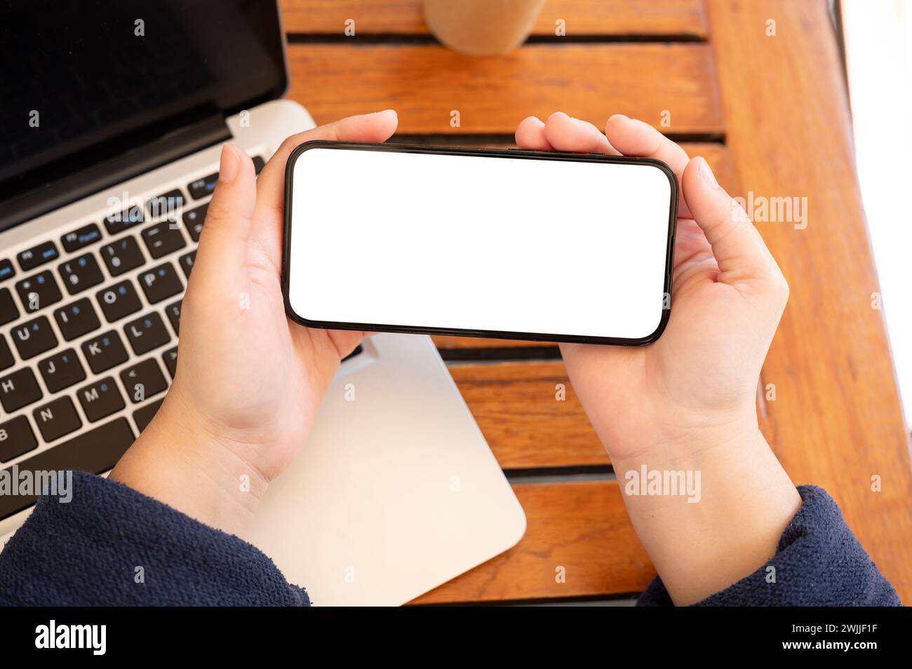 A top view image of a woman holding a white-screen smartphone mockup in a horizontal position, using her smartphone at a table indoors. Stock Photo
