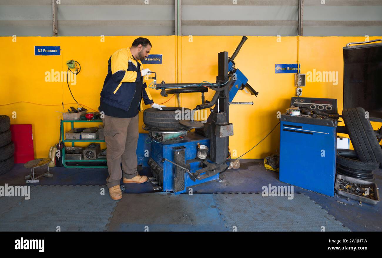 A technician in uniform and glove at an automotive service center working with a tire-changing machine. There are various garage equipment and tools i Stock Photo