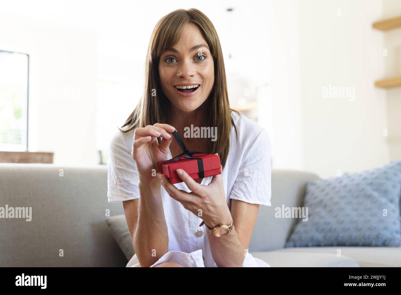 A young Caucasian woman appears delighted as she opens a small red gift box Stock Photo