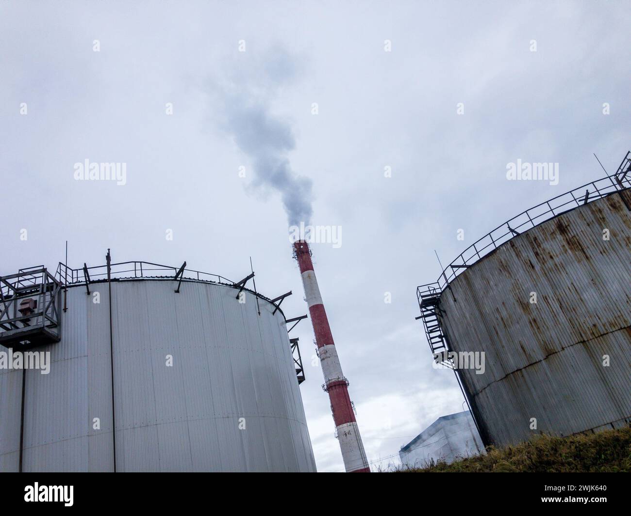 Industrial landscape with oil storage tanks and smokestack emitting fumes against a cloudy sky, implying environmental impact. Stock Photo