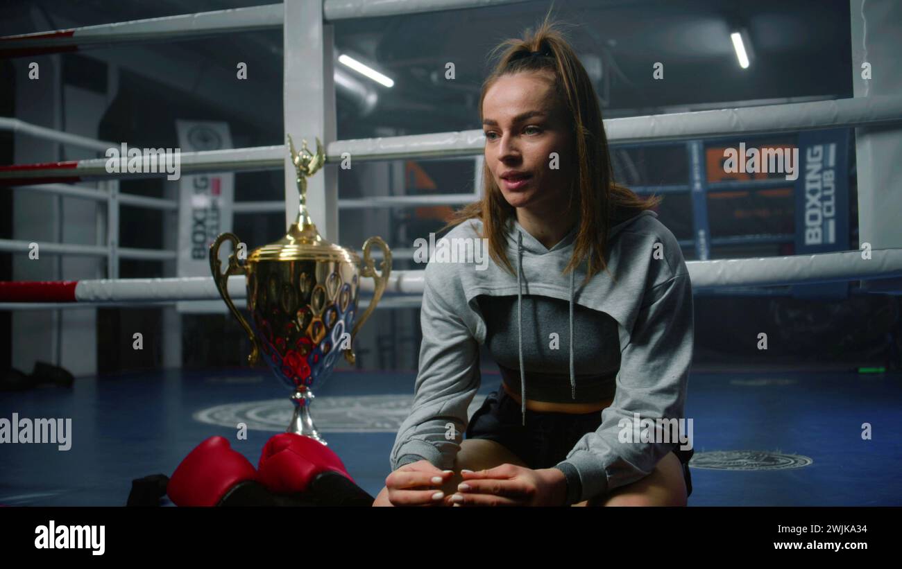 Female boxer sits at boxing ring and discusses victory in tournament with interviewer. Athlete talks after championship or competition in boxing gym. Cup of winner and boxing gloves lies near fighter. Stock Photo